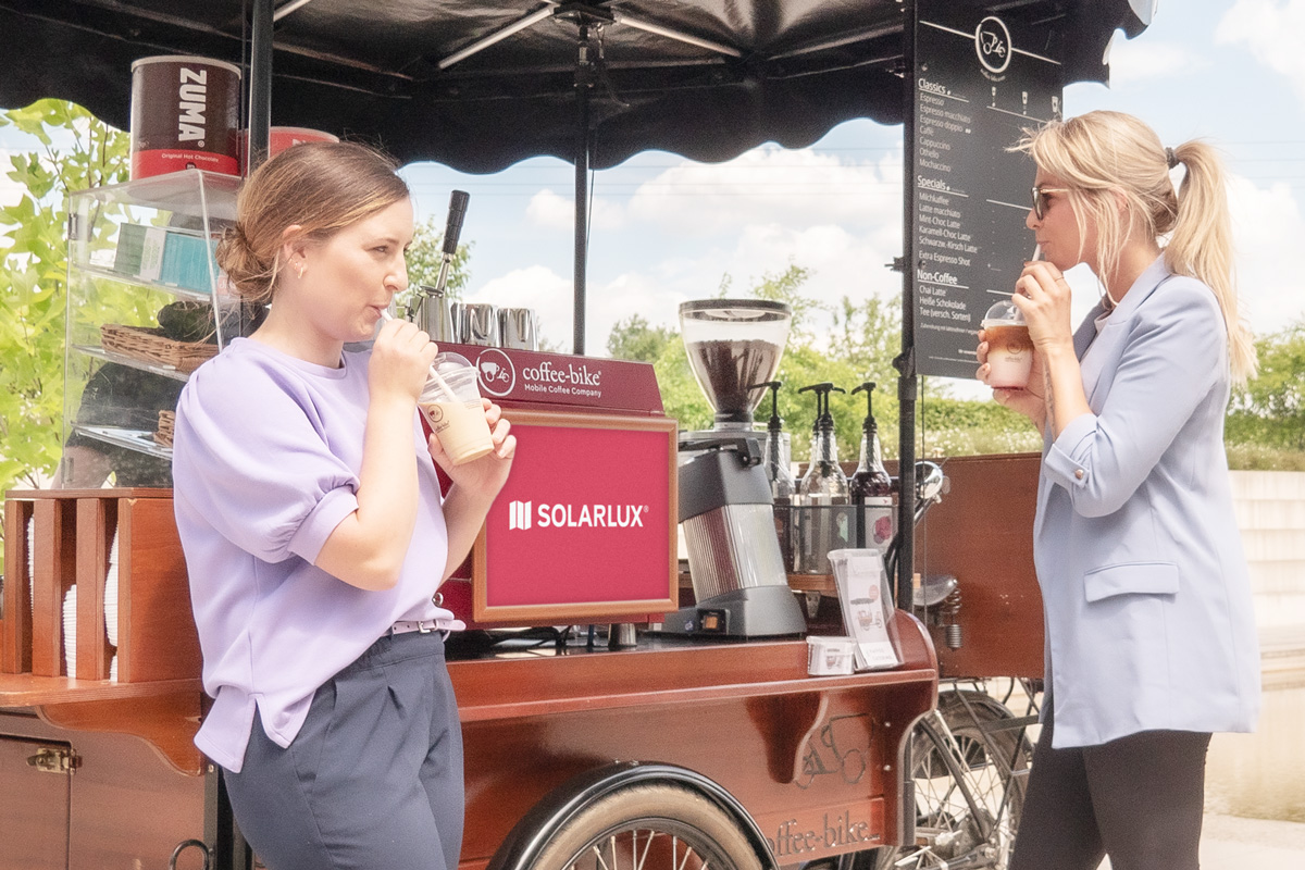 Zwei Frauen trinken Kaffee vor dem Coffee-Bike, das ein Solarlux Poster zeigt