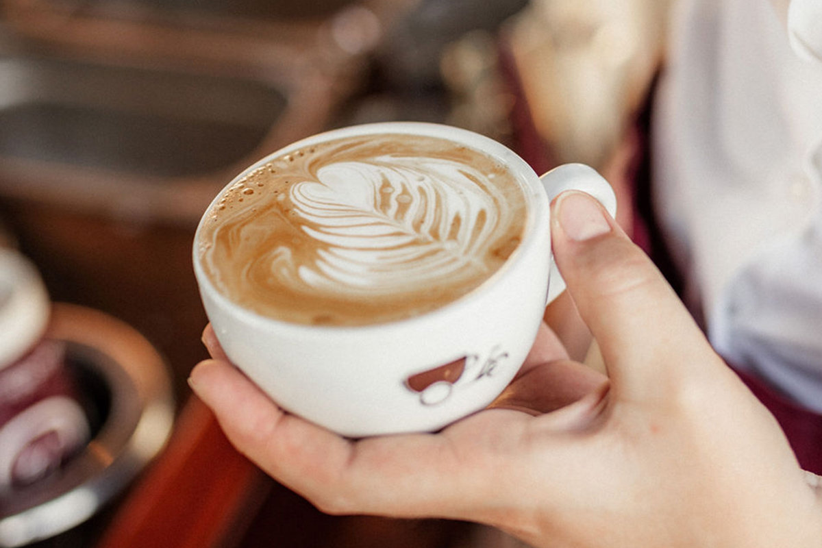 The hand of a barista holds a white porcelain Coffee-Bike cup with a latte art heart 