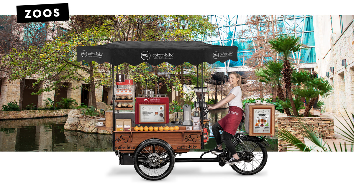 A barista in company clothes is sitting smiling on a coffee bike in side view against a background showing a zoo enclosure with the word zoos in the upper left corner
