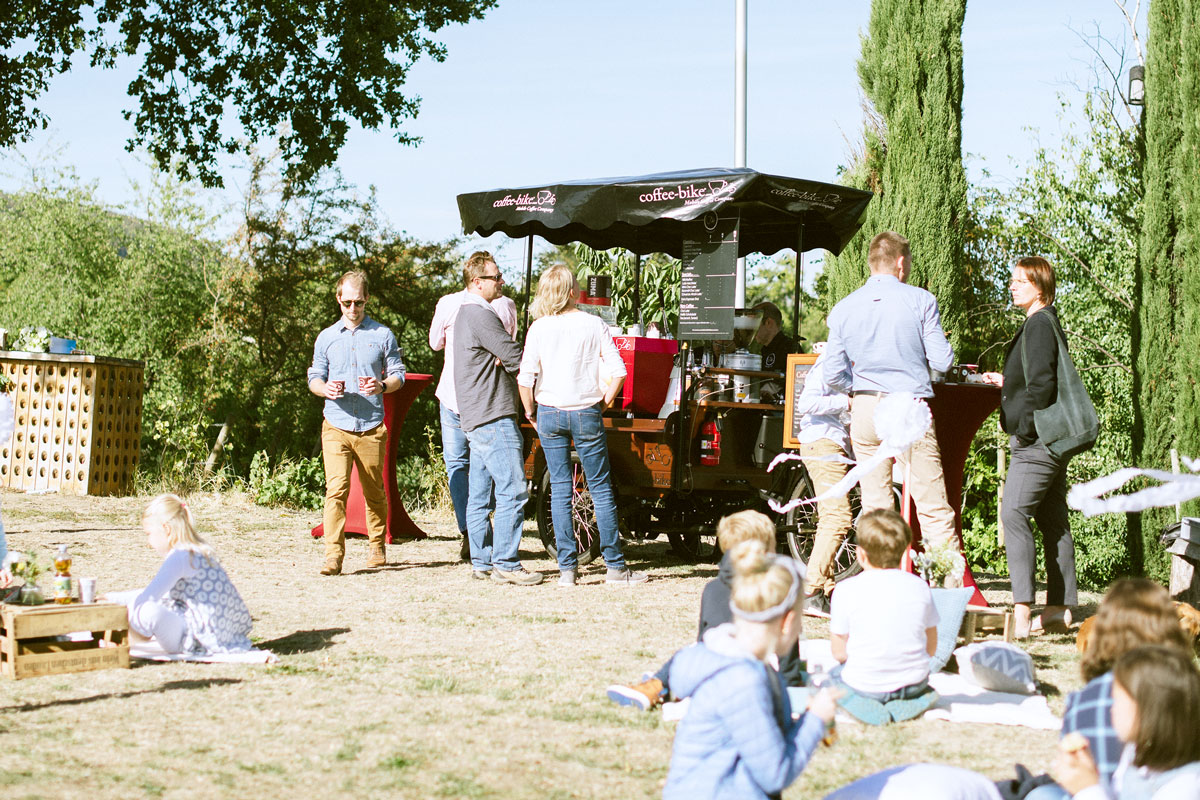 Children and adults stand and sit on a lawn where a barista on a Coffee-Bike serves specialty coffees